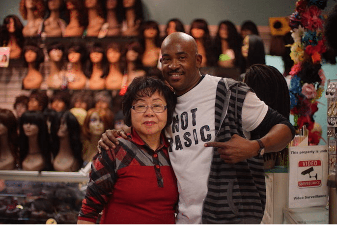 Hosa Anthony, an African-American man, embraces Soon Hong, an Asian woman, in front of a wall display of mannequins wearing wigs. Anthony, at right, is a wig stylist at Mondawmin Mall's Modern Wigs shop. He's been working at Modern Wigs for more than 20 years. Hong is the store manager and has been there for more than 40 years. The wig store relies on older customers who have been coming to the store for decades.